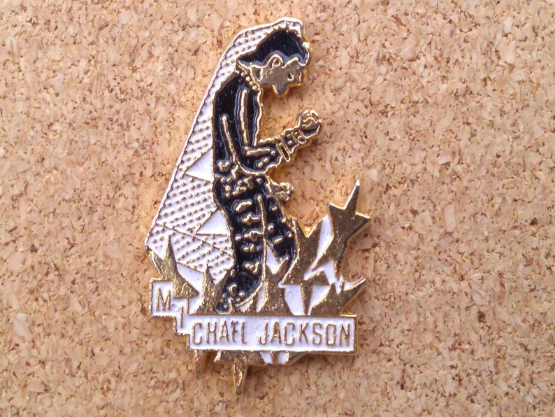 K&ouml;nnte beinhalten: Goldfarbene Anstecknadel mit einem schwarz-wei&szlig;en Cartoon-Bild von Michael Jackson in einem schwarzen Anzug und wei&szlig;em Handschuh, der auf einem wei&szlig;en und goldenen Stern kniet. Der Text "MICHAEL JACKSON" ist in wei&szlig;en Buchstaben auf einem goldenen Banner geschrieben.