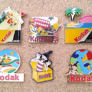 Puede incluir: Seis pines de esmalte coloridos que presentan una variedad de escenas y el logotipo de Kodak. Los pines presentan un esquiador, un autobús, una palmera, un castillo, una caja y un globo terráqueo.