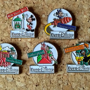 May include: Five Euro Disney pins featuring different themed lands. The pins are circular with a white background and feature a colourful illustration of the land's theme. The pins are all labelled with the land's name and "Euro Disney".