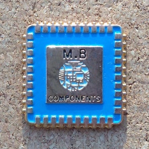 Op de afbeelding: Een gouden speld in de vorm van een computerchip met een blauwe achtergrond. De speld heeft de tekst "M.B. COMPONENTS" in witte letters.