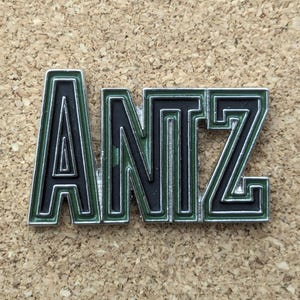 Puede incluir: Un emblema metálico con la palabra "ANTZ" en una fuente audaz y estilizada. Las letras son negras con un contorno verde y un borde plateado. El emblema está sobre un fondo texturizado y beige.