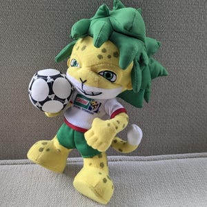 May include: A plush toy of a yellow jaguar-like mascot holding a soccer ball. The mascot has green hair, green shorts, and a white shirt with a sponsor logo. The soccer ball is black and white.