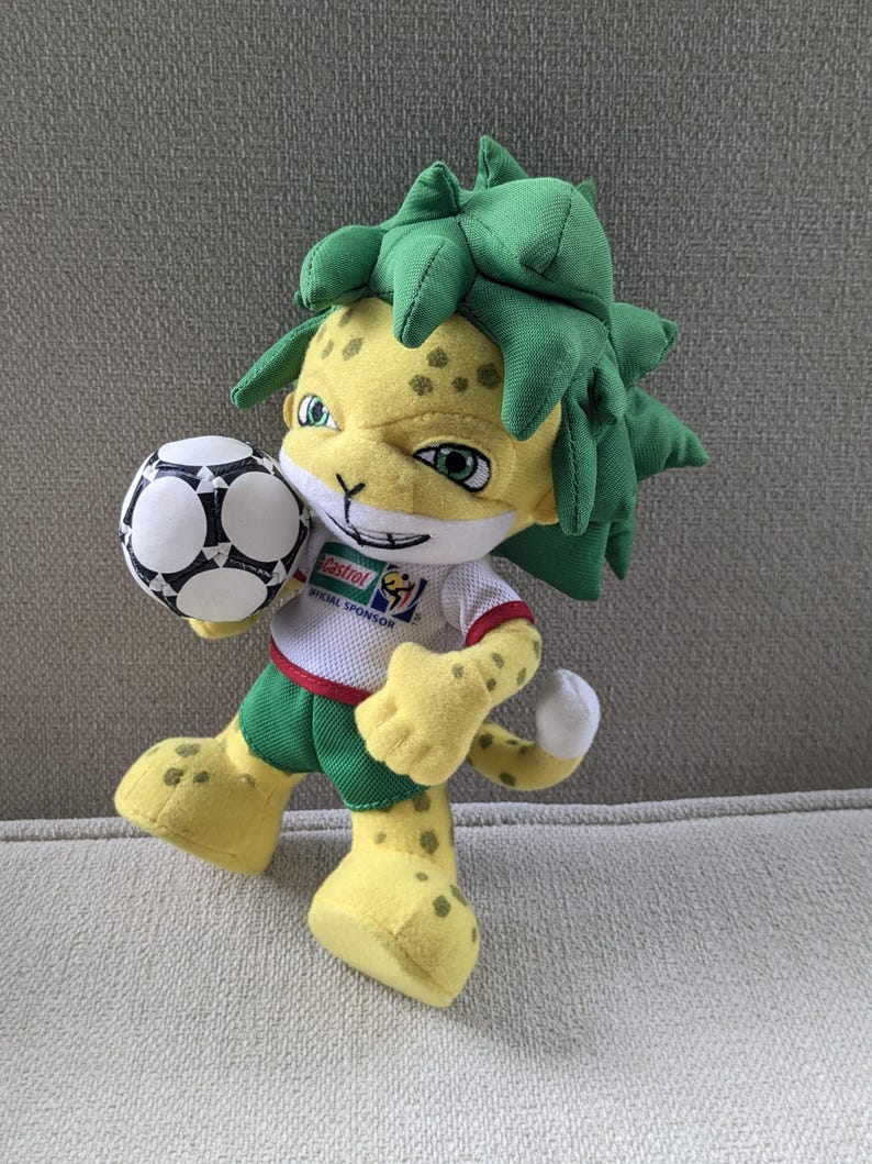 May include: A plush toy of a yellow jaguar-like mascot holding a soccer ball. The mascot has green hair, green shorts, and a white shirt with a sponsor logo. The soccer ball is black and white.
