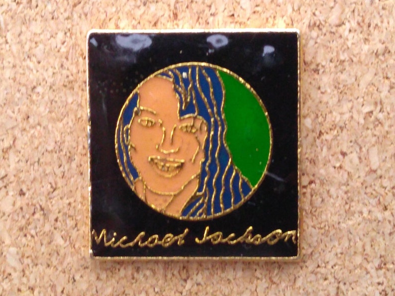 K&ouml;nnte beinhalten: Eine goldfarbene Anstecknadel mit schwarzem Hintergrund und einem runden Bild von Michael Jackson. Das Bild ist von einem blauen und gr&uuml;nen Rand umgeben. Der Text "Michael Jackson" ist in Gold unter dem Bild geschrieben.