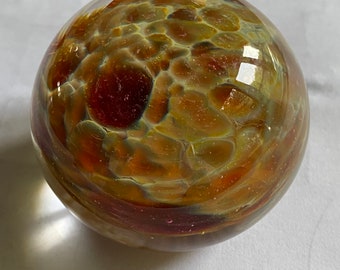 May include: A clear glass sphere with internal patterns in shades of brown, orange, and yellow. The sphere has a textured appearance, with a mottled design throughout. The glass reflects light, creating highlights and shadows.