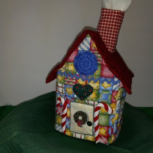 May include: A red and white patchwork fabric house-shaped tissue box cover with a candy cane design and a red velvet roof. The house has a wreath on the door and a chimney with a white fabric top.