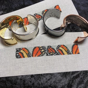 May include: Four metal cuff bracelets in gold, silver, and rose gold tones are displayed on a white canvas with a butterfly design. The canvas has the word "MONARCH" embroidered on it, with orange, yellow, and black butterfly wings.