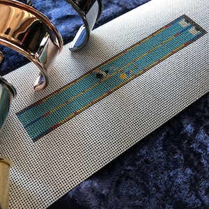 May include: A close-up of a needlepoint canvas with a blue and yellow design. The canvas is lying on a dark blue surface. There are several metal bracelets in the background.