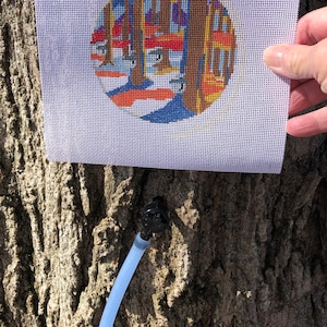 May include: A white canvas with a colorful circular design of a building with windows. The canvas is being held by a hand in front of a tree trunk.