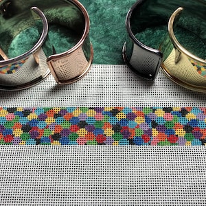 May include: Three metal bracelets with different finishes: silver, gold, and black. The bracelets are lying on a white canvas with a colorful patterned fabric strip in the center.