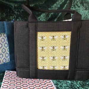 May include: Black tote bag with a yellow and white embroidered panel featuring a repeating pattern of bees. The panel is framed with a black border.