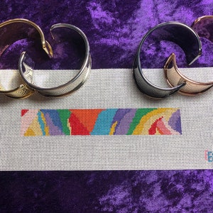 May include: Three metal bracelets in gold, silver, and black, arranged on a white canvas with a colorful striped design. The canvas is on a purple background.