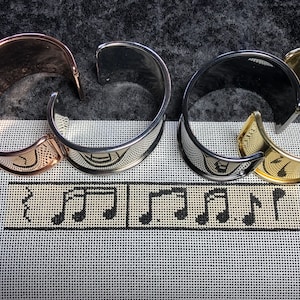 May include: Four metal bracelets in different colors: rose gold, silver, black, and gold. The bracelets are arranged in a row on a white canvas with black musical notes printed on it.