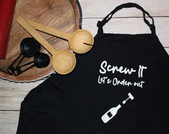 May include: Black kitchen apron with the text "Screw it Let's Order out" in white lettering. A white outline of a wine bottle with a corkscrew is below the text.