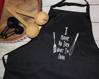 May include: Black kitchen apron with white text that reads "I have no idea what I'm doing" and a spatula and whisk graphic.