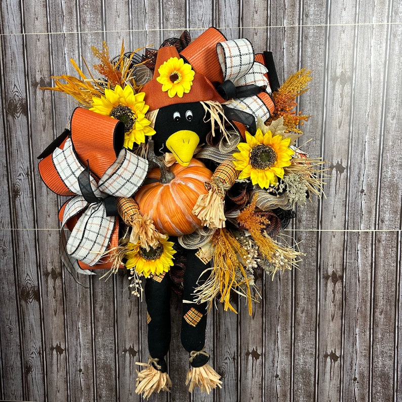 May include: A seasonal wreath with a scarecrow design. The wreath features a black crow face with a yellow beak, a pumpkin, sunflowers, and orange and plaid ribbons. The scarecrow has black legs with plaid patches and straw feet.