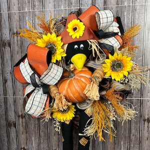 May include: A seasonal wreath with a scarecrow theme. It features a black crow head with a yellow beak, a pumpkin, sunflowers, and orange and plaid ribbons. The wreath is decorated with dried wheat and other autumn foliage.