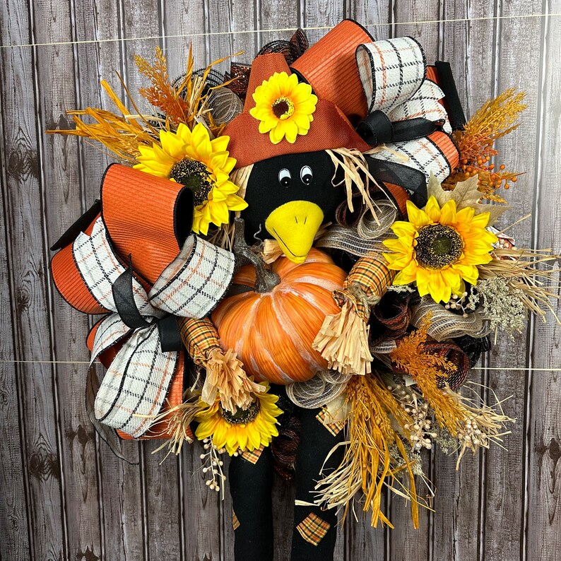 May include: A seasonal wreath with a scarecrow theme. It features a black crow head with a yellow beak, a pumpkin, sunflowers, and orange and plaid ribbons. The wreath is decorated with dried wheat and other autumn foliage.