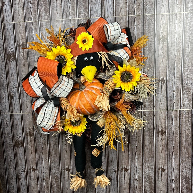 May include: A seasonal wreath with a scarecrow design. The wreath incorporates a black crow head with a yellow beak, a pumpkin, sunflowers, and orange and plaid ribbons. The scarecrow has black legs with straw feet.