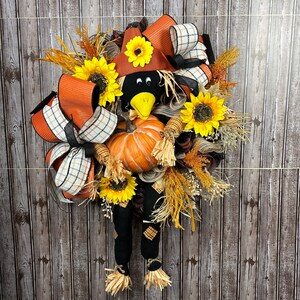 May include: A seasonal wreath with a scarecrow design. It features a pumpkin, sunflowers, and orange, black, and white ribbons. The scarecrow has a black head and legs, a brown hat, and a yellow beak. The wreath is decorated with autumnal elements.