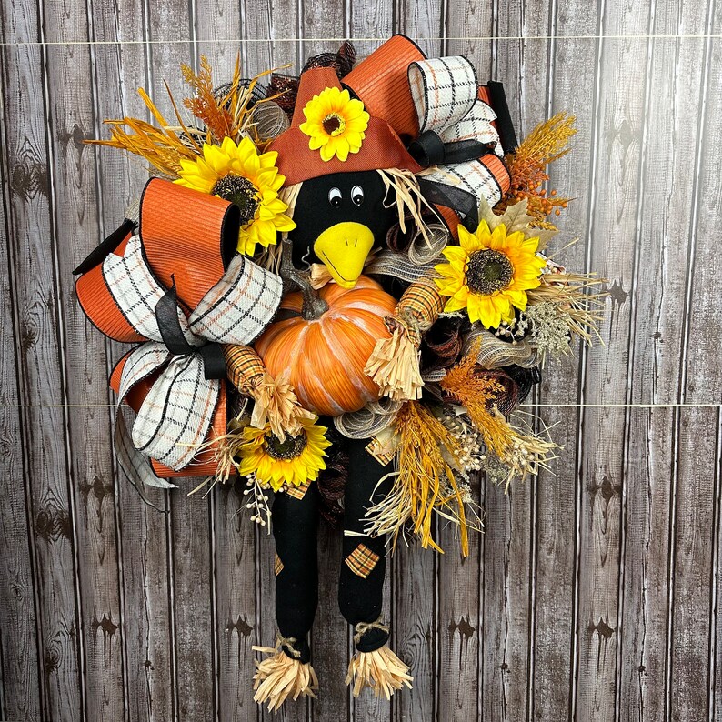 May include: A seasonal wreath with a scarecrow design. It features a pumpkin, sunflowers, and orange, black, and white ribbons. The scarecrow has a black head and legs, a brown hat, and a yellow beak. The wreath is decorated with autumnal elements.