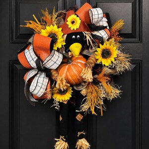May include: A decorative autumn wreath with a scarecrow theme. The wreath features a black crow head, a pumpkin, sunflowers, and orange and white plaid ribbons. It is decorated with dried grasses and a brown hat, perfect for the season.