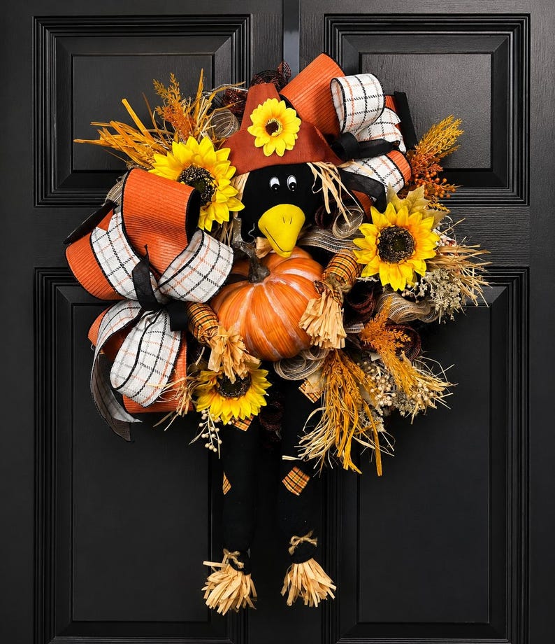 May include: A decorative autumn wreath with a scarecrow theme. The wreath features a black crow head, a pumpkin, sunflowers, and orange and white plaid ribbons. It is decorated with dried grasses and a brown hat, perfect for the season.