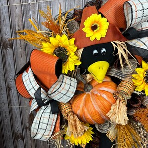 May include: A seasonal wreath with a crow, pumpkin, sunflowers, and ribbons. The crow wears a brown hat with a sunflower. The wreath incorporates orange, black, and white plaid ribbons, and dried wheat, creating a harvest theme.
