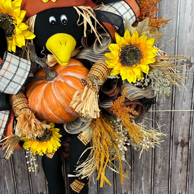May include: A seasonal wreath with a black crow figure, a yellow beak, and eyes, an orange pumpkin, and sunflowers. The wreath is adorned with plaid ribbons, dried wheat, and autumn leaves, creating a rustic and festive appearance.