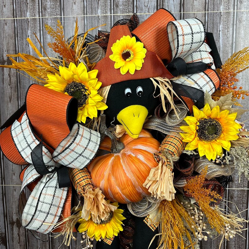 May include: A seasonal wreath with a crow figure, pumpkins, and sunflowers. The crow has a black head, a yellow beak, and a brown hat. The wreath is decorated with orange and plaid ribbons, dried grasses, and fall foliage. The wreath is a festive autumn decoration.