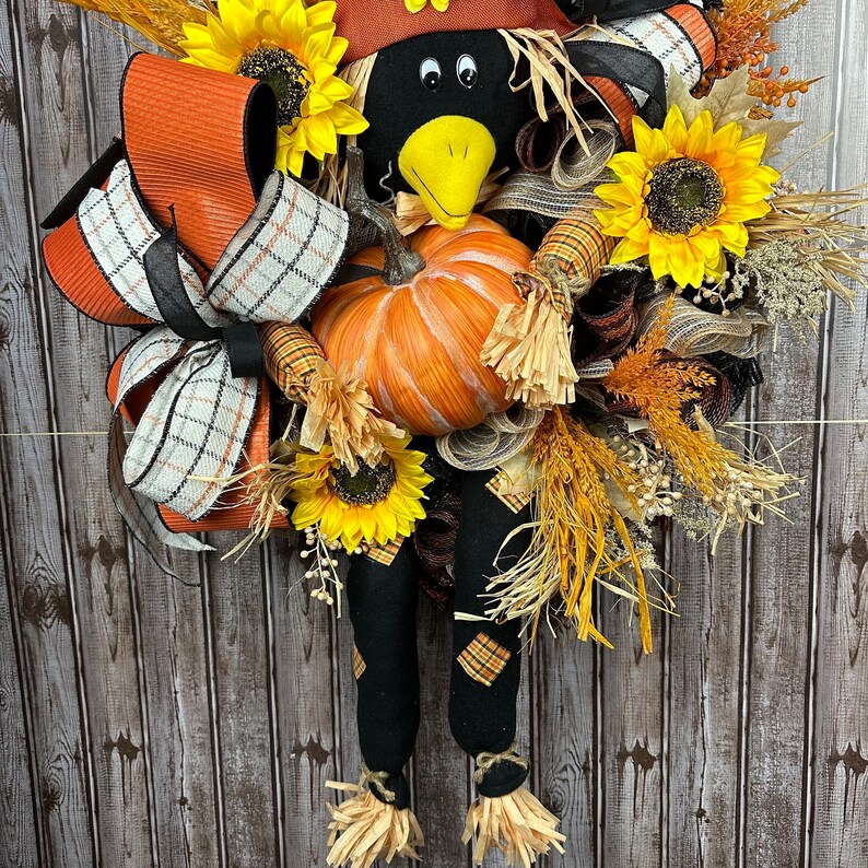 May include: A handmade scarecrow wreath featuring a black crow head, a pumpkin, and sunflowers. The wreath incorporates orange, black, and white plaid ribbons, and straw accents. The scarecrow's legs are black with plaid patches.