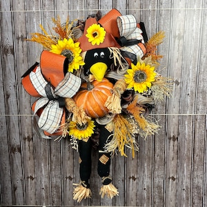 May include: A seasonal wreath with a scarecrow design. The wreath features a black crow head with a brown hat, a pumpkin, sunflowers, and orange and plaid ribbons. The scarecrow has black legs with plaid patches and straw feet.
