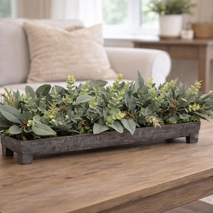May include: A rectangular, weathered gray wooden planter box filled with faux greenery, including various shades of green leaves and sprigs. The planter sits on a wooden coffee table. The background includes a beige sofa and a window.