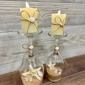 May include: Two glass candle holders with a nautical theme. Each holder has a white pillar candle, a seashell, and a starfish inside. The holders are decorated with twine and a bow.