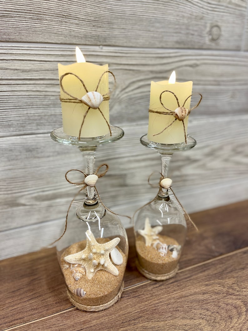 May include: Two glass candle holders with a nautical theme. Each holder has a white pillar candle, a seashell, and a starfish inside. The holders are decorated with twine and a bow.