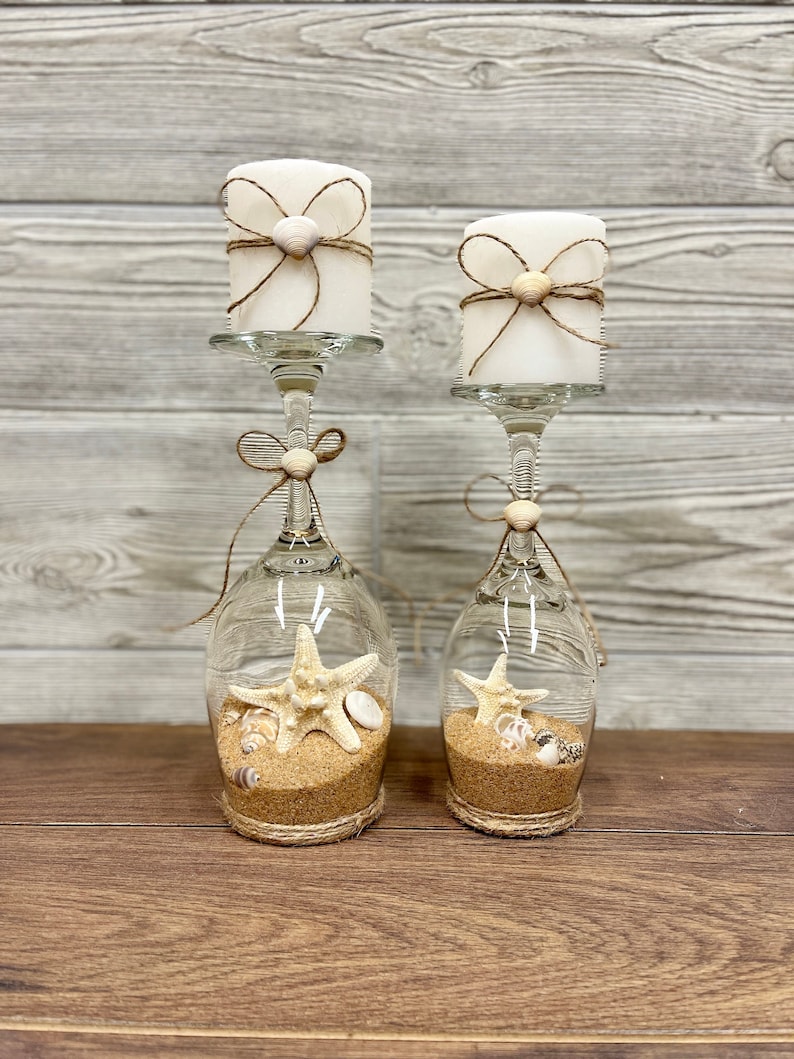 May include: Two glass candle holders with white candles, decorated with twine and seashells. The holders are filled with sand and seashells, including a starfish.