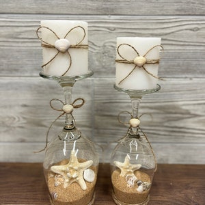 May include: Two white pillar candles with twine and seashell accents are displayed in glass wine goblets filled with sand and seashells. The goblets are tied with twine at the base. The text "Wax Candles" is displayed above the candles.