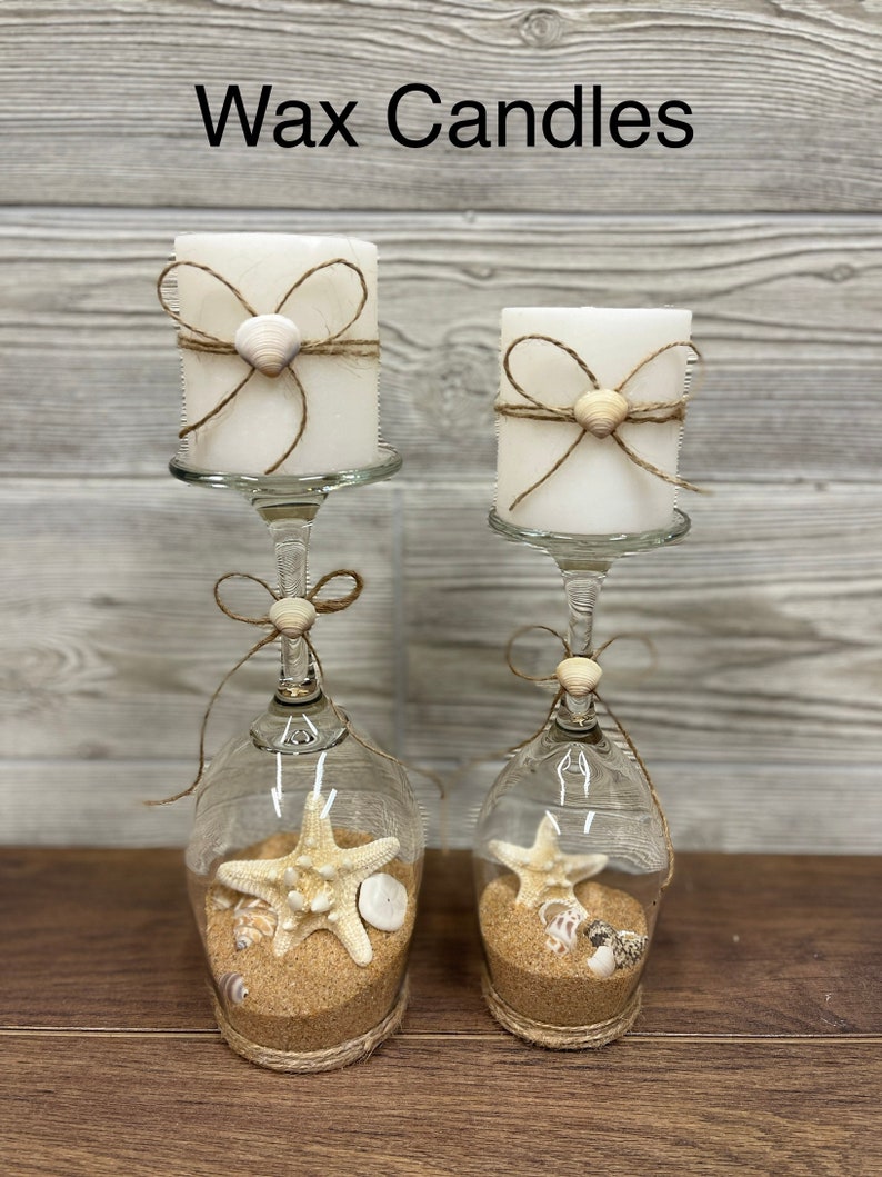May include: Two white pillar candles with twine and seashell accents are displayed in glass wine goblets filled with sand and seashells. The goblets are tied with twine at the base. The text "Wax Candles" is displayed above the candles.