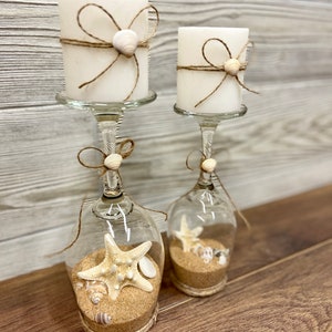 May include: Two white candles with twine and seashells wrapped around them are placed in clear glass wine glasses with sand and seashells inside. The glasses are on a wooden surface.