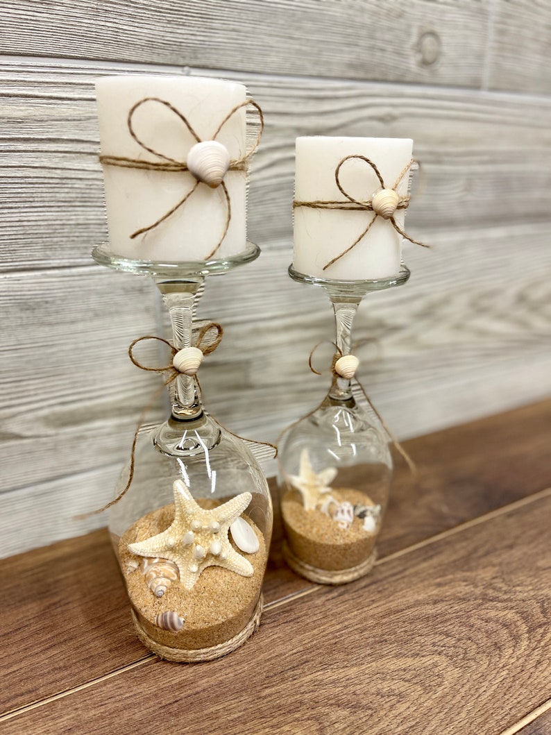 May include: Two white candles with twine and seashells wrapped around them are placed in clear glass wine glasses with sand and seashells inside. The glasses are on a wooden surface.