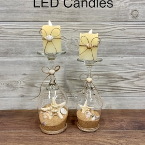 May include: Two LED candles in glass candle holders with a seashell and starfish design. The candles are wrapped in twine and have a seashell accent. The candle holders are filled with sand and seashells.