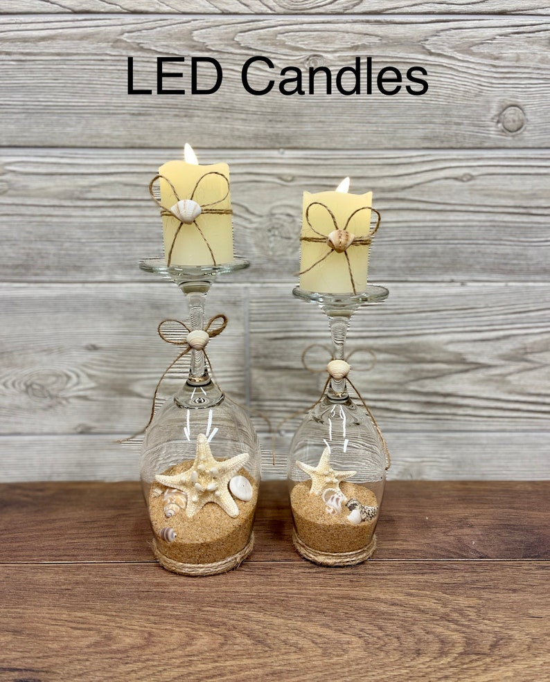 May include: Two LED candles in glass candle holders with a seashell and starfish design. The candles are wrapped in twine and have a seashell accent. The candle holders are filled with sand and seashells.