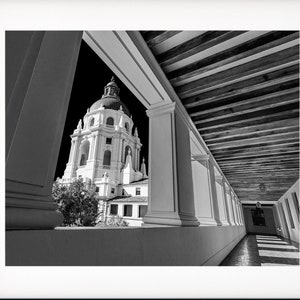 May include: A black and white photograph of a building with a dome, seen through a colonnade. The building is framed by the columns and the wooden ceiling of the colonnade.