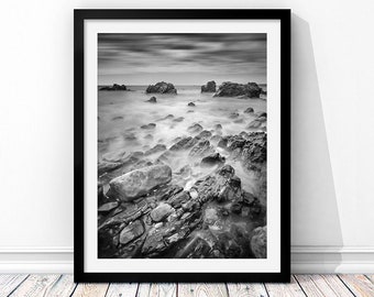May include: Black and white photograph of a rocky coastline with the ocean in the background. The water is blurred, creating a sense of motion. The rocks are in the foreground, and the sky is cloudy.