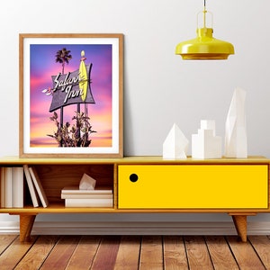 K&ouml;nnte beinhalten: Ein gerahmtes Foto eines Vintage-Motelschildes mit den Worten "Safari Inn" in Neonlicht. Das Schild ist vor einem farbenfrohen Sonnenuntergang mit Palmen im Hintergrund beleuchtet.