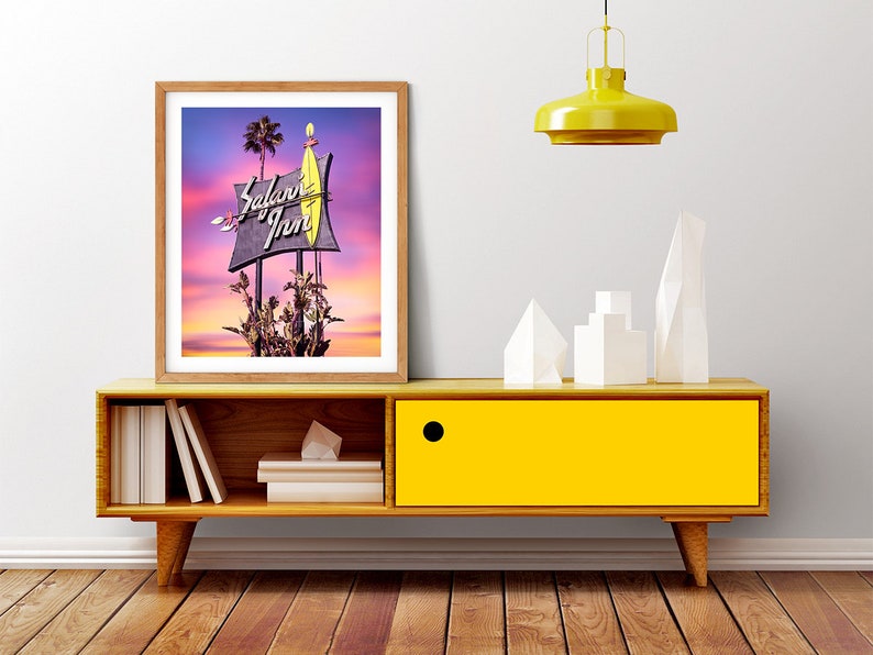 K&ouml;nnte beinhalten: Ein gerahmtes Foto eines Vintage-Motelschildes mit den Worten "Safari Inn" in Neonlicht. Das Schild ist vor einem farbenfrohen Sonnenuntergang mit Palmen im Hintergrund beleuchtet.