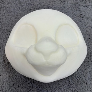 May include: A blank, off-white fursuit head base, featuring eye openings and a prominent snout. The head is smooth and rounded, designed for customization. The base is made of a lightweight material.