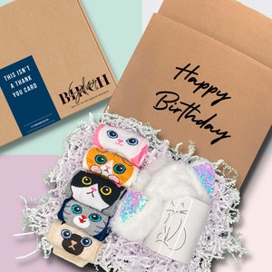 May include: A brown cardboard box with the words "Happy Birthday" written on it. The box is filled with white shredded paper and contains a white mug with a cat design, a white headband with iridescent sequins, and five pairs of cat-themed socks.