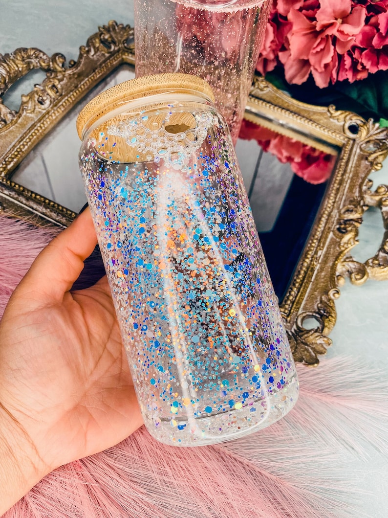 Peut inclure: Une bouteille en verre transparente avec un couvercle en bois remplie de paillettes bleues et argent&eacute;es iris&eacute;es. La bouteille est tenue dans une main.