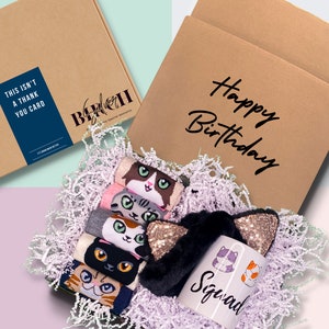 May include: A brown cardboard box with the text "Happy Birthday" on the front. The box is filled with white tissue paper and contains a cat-themed gift set. The gift set includes a mug with the text "Squad" and a cat design, a black headband with gold glitter cat ears, and a set of cat-themed socks.